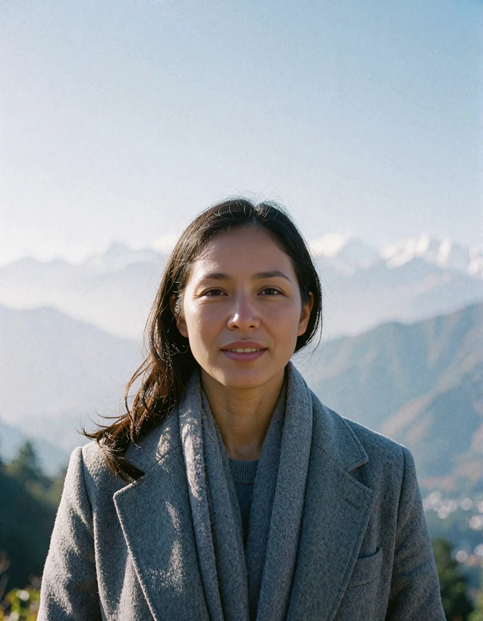 Nepali member in a stone-grey wool coat outdoors with distant Himalayan foothills behind her