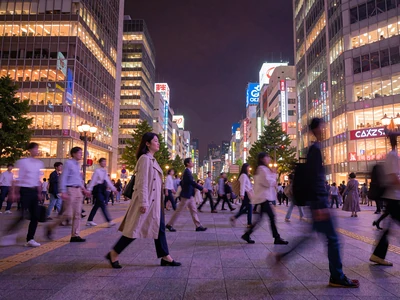 Japanese woman in a light trench coat walking through a busy illuminated Tokyo crossing at night