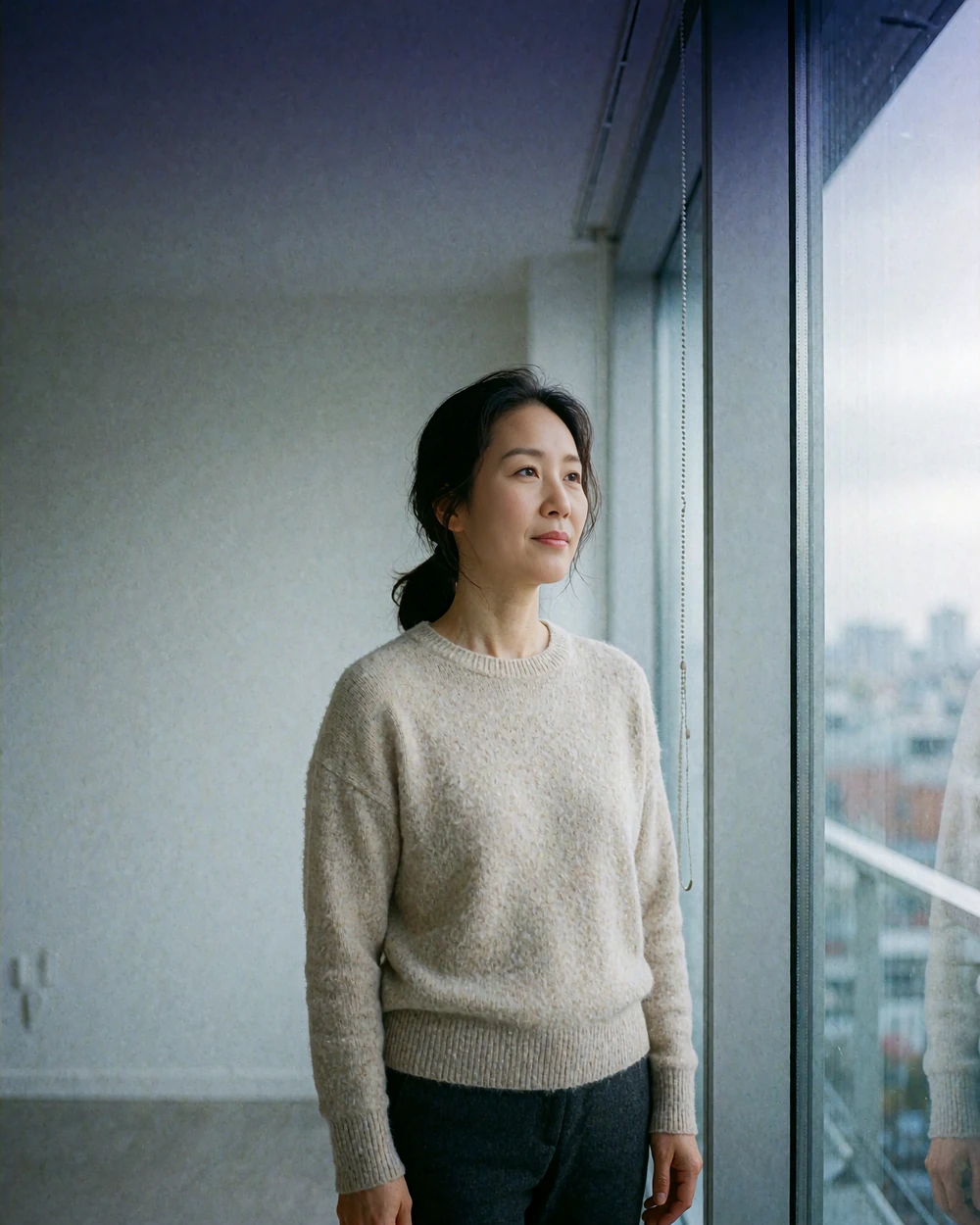 Thoughtful Korean woman standing beside a window in a calm modern apartment